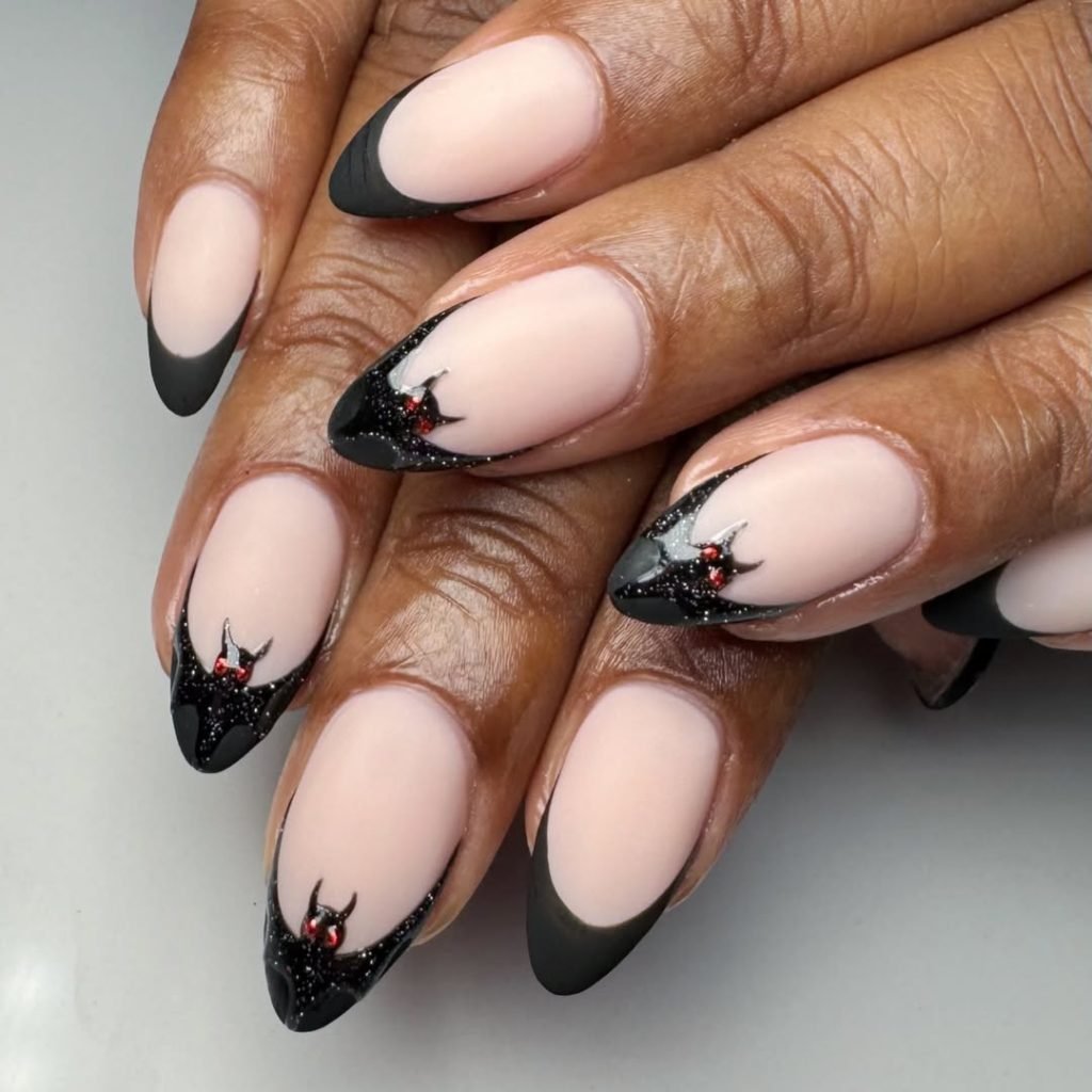 Matte nude almond nails with black French tips and 3D black glitter bats with red gem eyes, a wearable black Halloween nail idea.