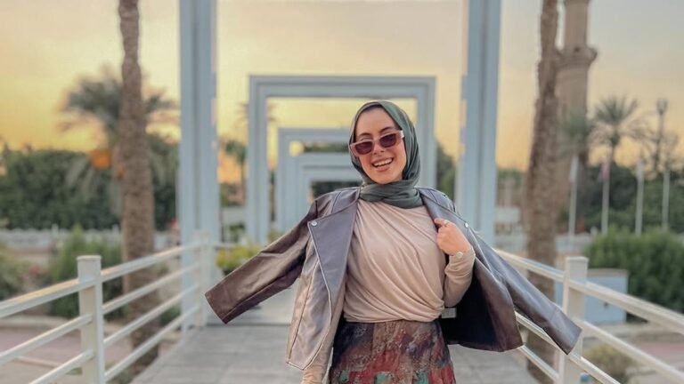 Woman in a hijab twirling on a sunset walkway lined with archways, wearing a brown leather jacket, beige top, and a colorful patchwork maxi skirt with brown platform boots