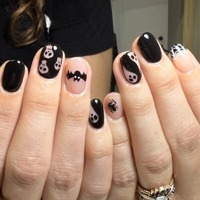 Short glossy black nails with a nude accent nail featuring a cartoon ghost, skull-and-crossbones nail art, and a spiderweb tip for a chic Halloween manicure