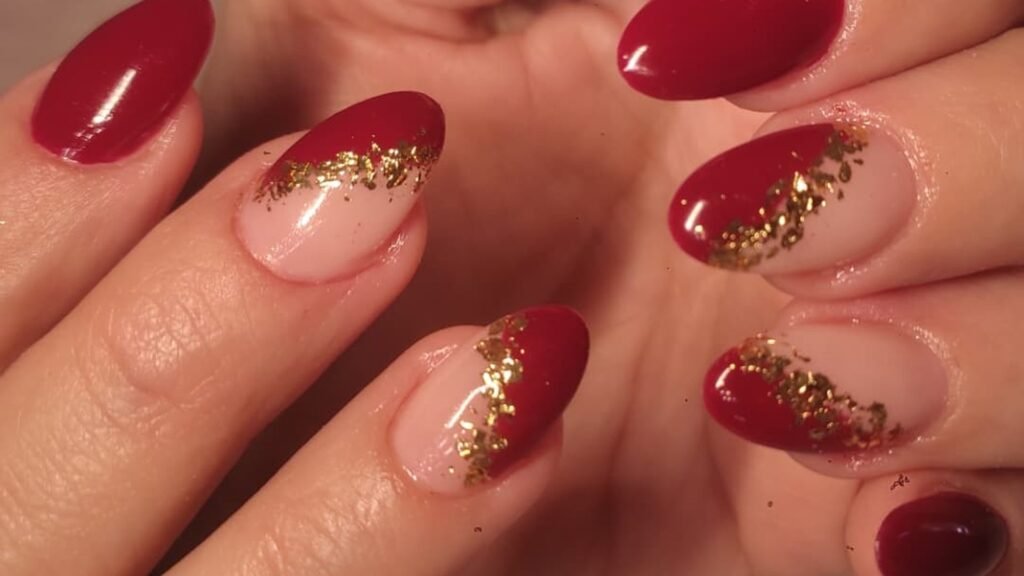 Close-up of glossy deep red almond nails, four of them split diagonally between red and sheer nude with a streak of gold foil along the line, while the remaining nails stay solid red.