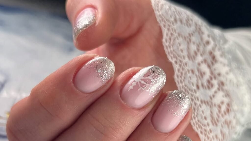 Close-up of a hand with short blush pink nails fading into silver glitter tips, one nail with a white hand-painted snowflake, resting against a white lace sleeve cuff.