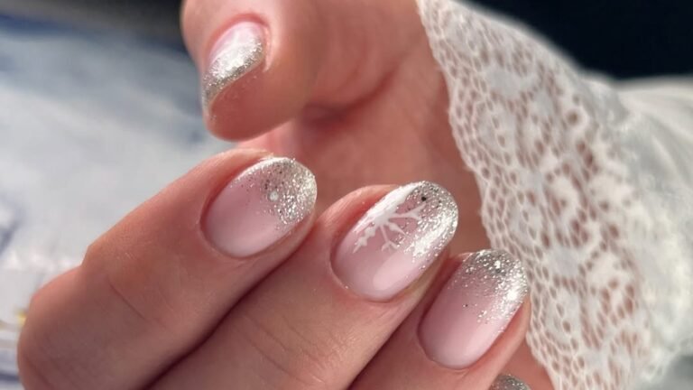 Close-up of a hand with short blush pink nails fading into silver glitter tips, one nail with a white hand-painted snowflake, resting against a white lace sleeve cuff.