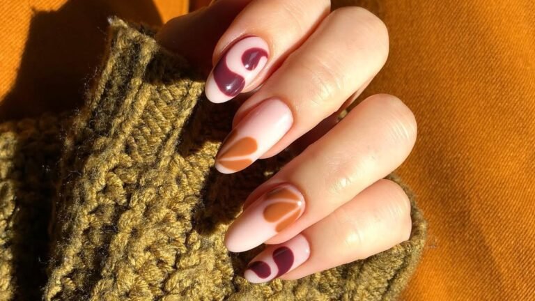 Almond gel nails painted with warm rust and oxblood autumn swirls, held against a mustard-yellow knit sweater
