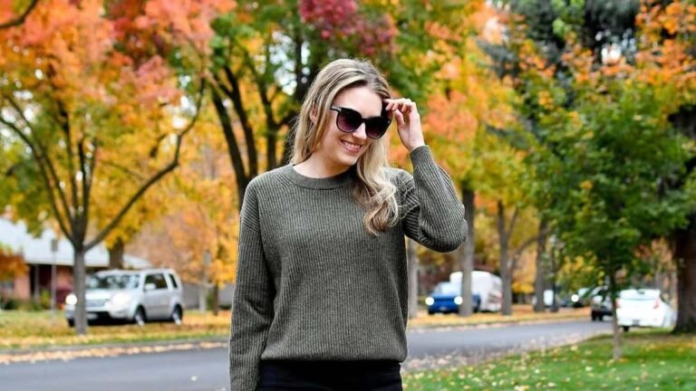 Woman in an olive green ribbed sweater and black skinny jeans carrying a burgundy handbag, standing on a street lined with vibrant red and orange autumn trees