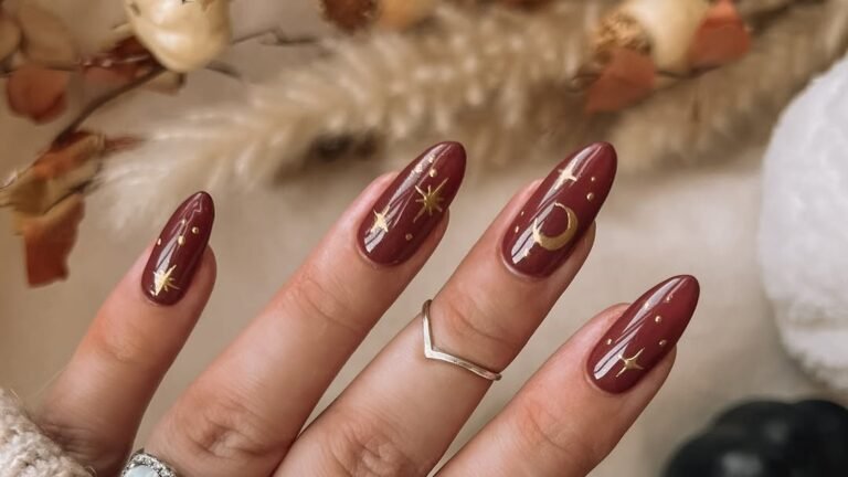 A hand with burgundy nails hand-painted with gold stars and a crescent moon rests near dried mini pumpkins and autumn leaves in a beige sweater cuff