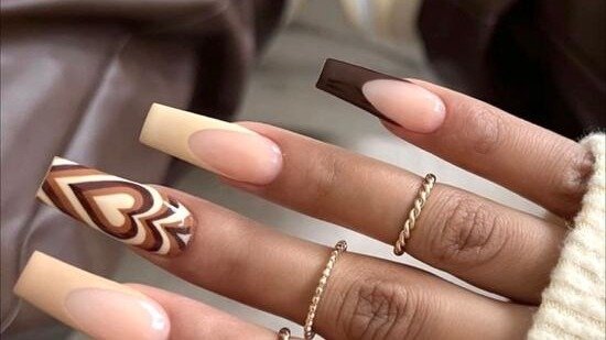Chocolate brown swirl heart nail art with nude and espresso accent nails, gold rings, on a warm brown leather background