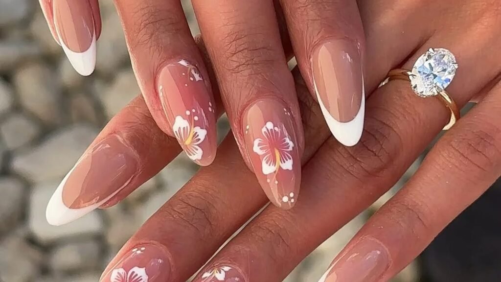 Almond nails with crisp white French tips and hand-painted white hibiscus flowers on a sheer pink base, one hand resting on the other beside an oval diamond ring on a gold band, with blurred gray stones behind.
