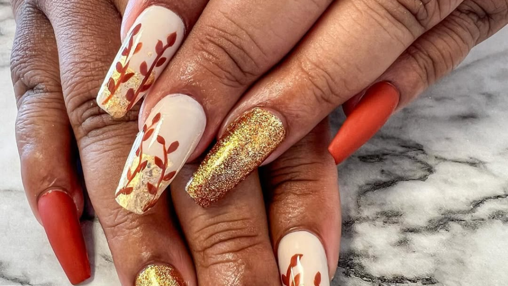 Close-up of two hands with long coffin-shaped Thanksgiving nails, featuring cream nails with gold foil vine and leaf art, one solid gold glitter accent nail, and matte rust nails, set against a white and gray marble background