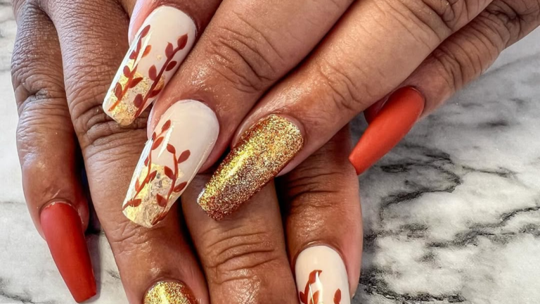 Close-up of two hands with long coffin-shaped Thanksgiving nails, featuring cream nails with gold foil vine and leaf art, one solid gold glitter accent nail, and matte rust nails, set against a white and gray marble background