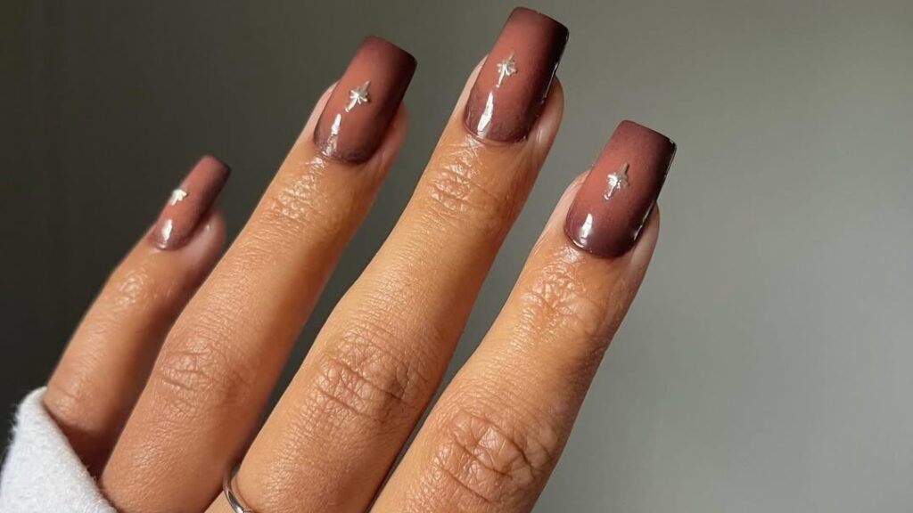 Four fingernails painted in matte deep mauve-brown, each with a tiny silver star charm placed near the cuticle, against a plain gray background