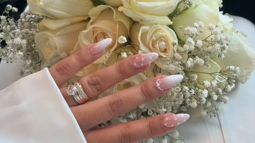 Hand with pink to white ombre almond nails decorated with tiny pearls and stacked diamond and gold rings, resting on a bouquet of cream roses and baby's breath beside a white satin sleeve.