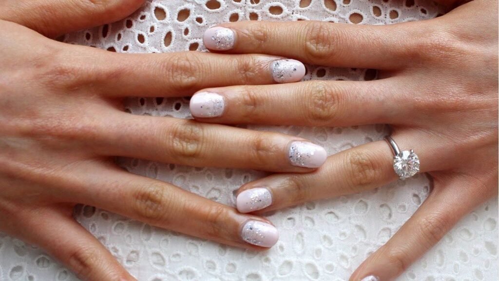 Two hands with short round blush pink nails dusted with silver glitter flecks resting across each other on white eyelet lace fabric, with a round solitaire diamond engagement ring on one finger.