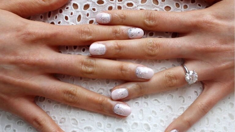 Two hands with short round blush pink nails dusted with silver glitter flecks resting across each other on white eyelet lace fabric, with a round solitaire diamond engagement ring on one finger.
