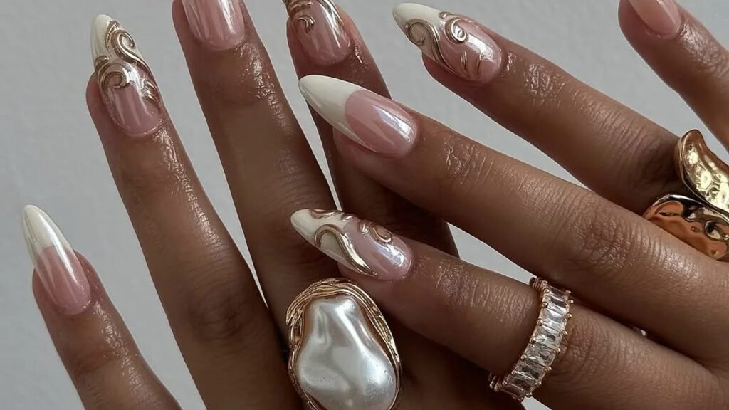 Almond nails with soft white French tips and raised gold swirl details on a sheer pink base, worn with a large baroque pearl ring, a gold band, and a crystal baguette ring.