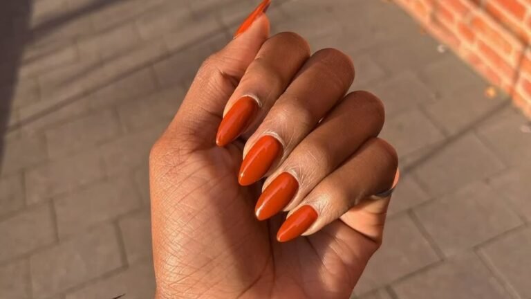 A hand with almond-shaped burnt orange nails catching sunlight, fingers curled against a blurred brick wall and paved sidewalk