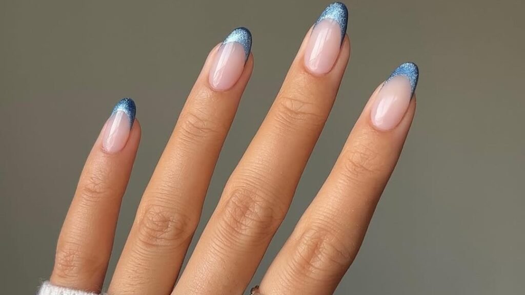 Nude nail bed with sparkling blue glitter French tips on almond-shaped nails, worn with a gold chain ring and cream sweater cuff