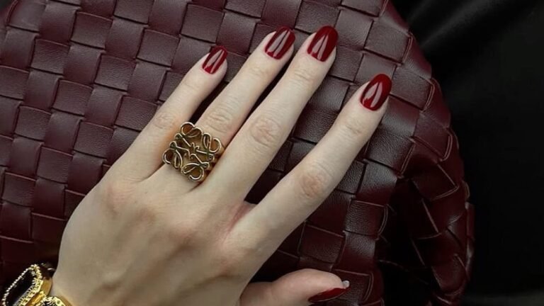 Woman's hand with deep oxblood red old money manicure resting on a woven burgundy leather bag with gold jewelry