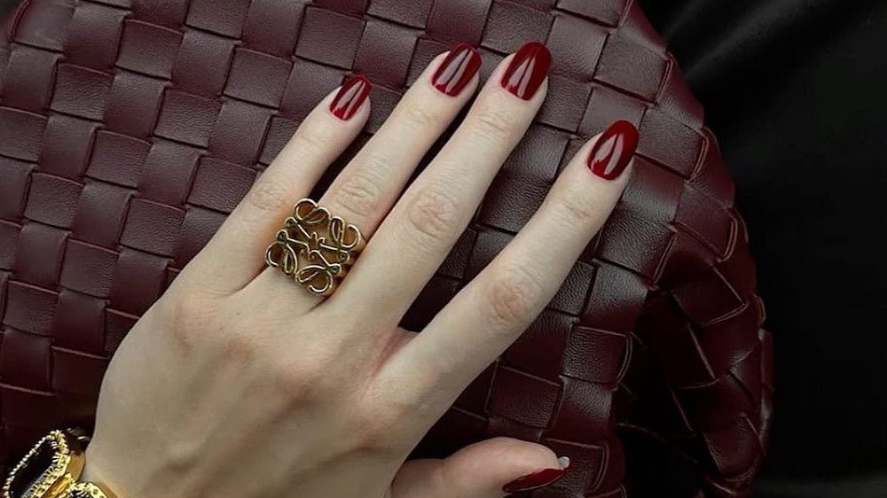 Woman's hand with deep oxblood red old money manicure resting on a woven burgundy leather bag with gold jewelry