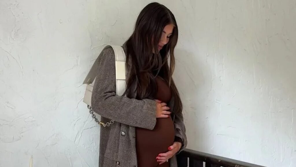 Pregnant woman in a fitted brown bodysuit and long herringbone coat standing on a staircase, showing a winter baby bump outfit with a bump-skimming base and a warm layer on top.