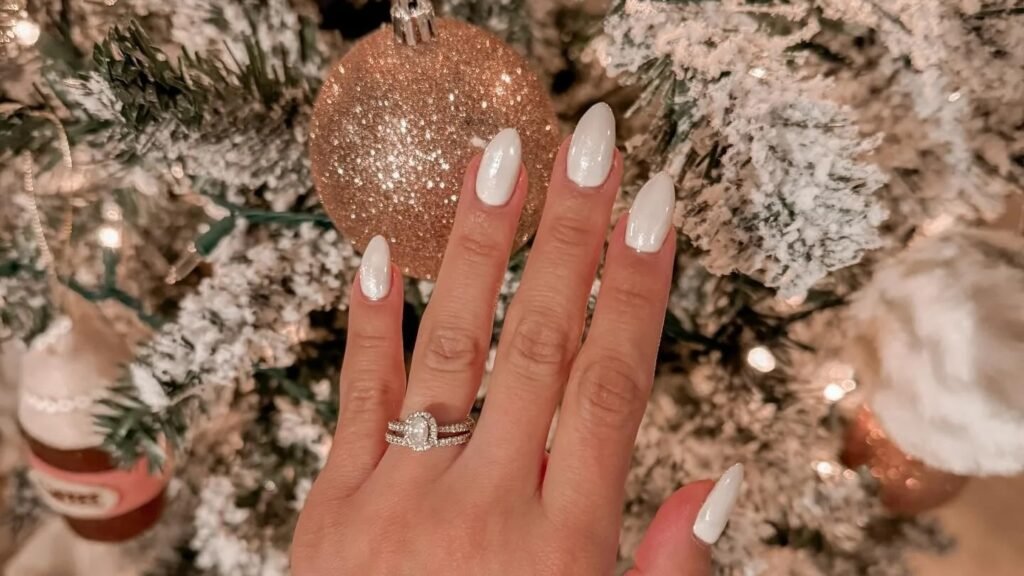 Glossy white manicure on oval nails resting near a gold glitter Christmas ornament on a snow flocked tree