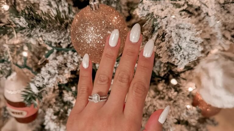 Glossy white manicure on oval nails resting near a gold glitter Christmas ornament on a snow flocked tree