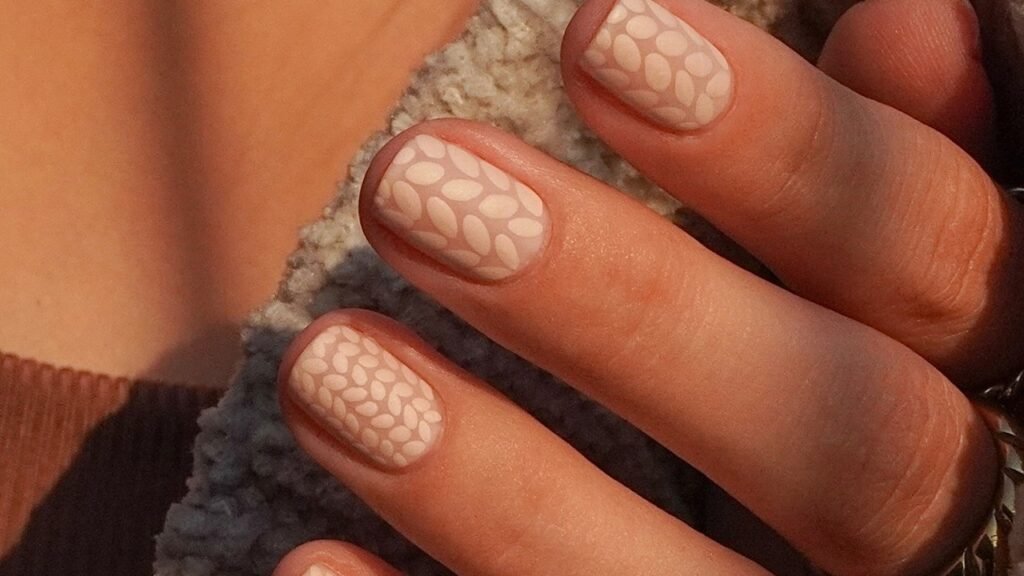 Nude nails with a raised leaf-stitch knit texture resting against a cream knit sweater sleeve