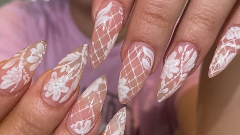 Close-up of long stiletto nails with hand-painted white lace lattice and raised floral patterns over a sheer nude pink base, with tiny pearl accents and the natural nail color showing through.