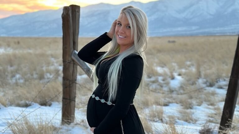 Pregnant woman smiling in a plunging black long-sleeve maternity dress with a concho belt and thigh slit, standing by a snowy fence line at sunset with mountains in the background