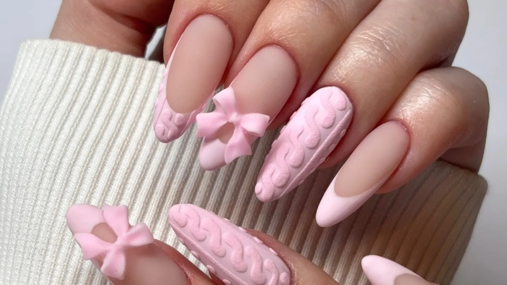 Pink cable-knit textured nails with a sculpted bow accent, styled with a matching sweater sleeve