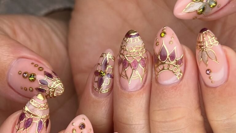 Close-up of almond-shaped nails painted sheer nude pink and decorated with thin gold line art, plum petal shapes, and tiny gold and green jewels, styled as a bridesmaid nail design.