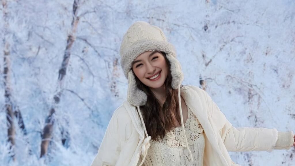 Smiling woman in a cream knit dress and puffer coat twirling in a snowy forest with frosted trees, one of 13 winter dress ideas