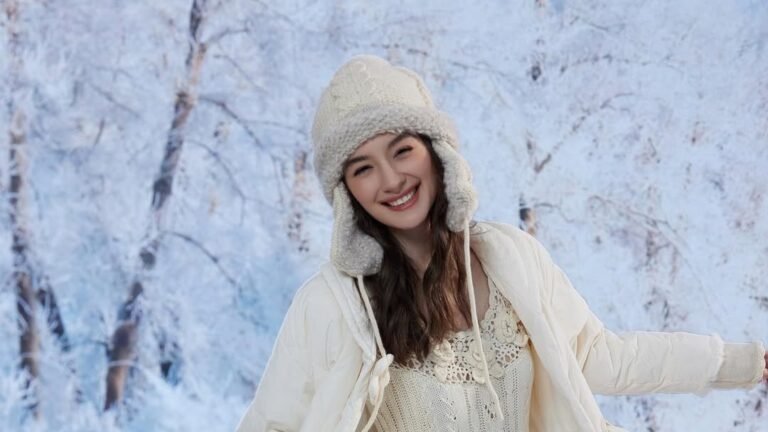 Smiling woman in a cream knit dress and puffer coat twirling in a snowy forest with frosted trees, one of 13 winter dress ideas