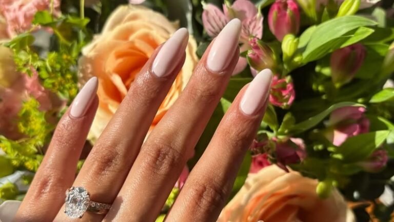 Hand with long milky blush almond-shaped nails and an oval diamond engagement ring held against a bouquet of peach roses, pink blooms, and green leaves in warm sunlight.