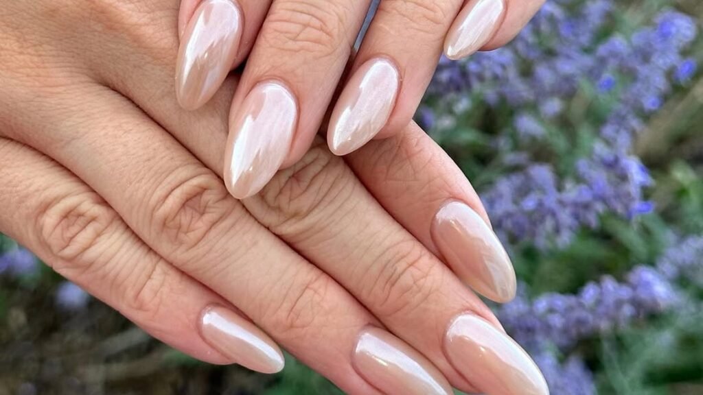 Close view of soft pearl chrome almond nails on layered hands with a thin silver ring band, set against purple lavender flowers in soft focus.