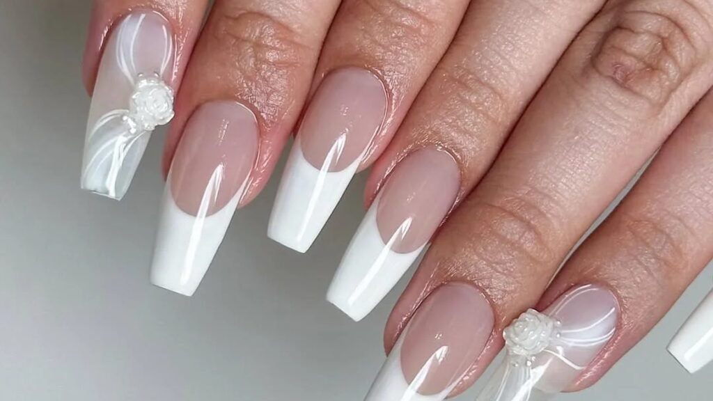 Long coffin nails with crisp white French tips over a sheer blush base, plus two accent nails topped with a small white pearl rose and soft white drape lines, shown against a light gray background.