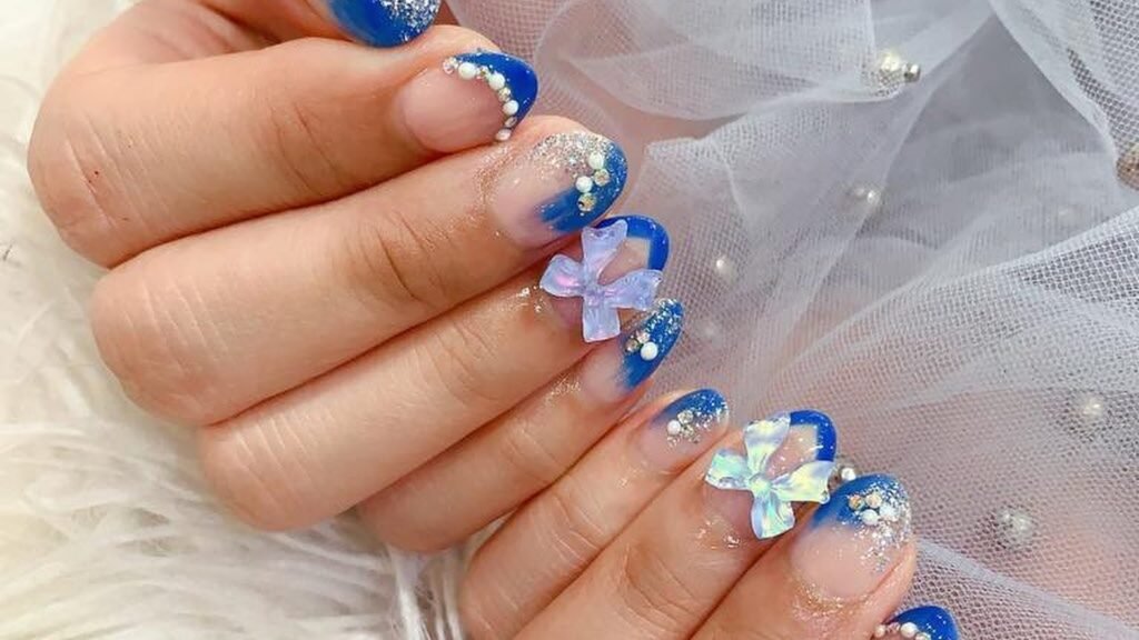 Blue wedding nails with royal blue glitter French tips, pearls, and two iridescent bows resting on white pearl-studded tulle and a fluffy white fur backdrop.