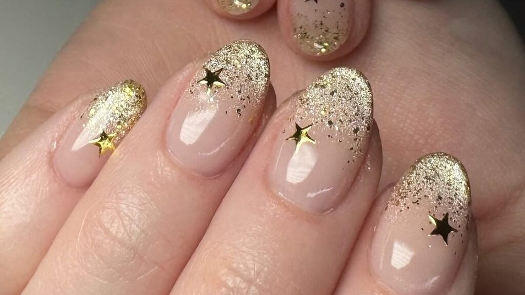 Glossy short oval nails with a sheer nude base, gold glitter fading up from the tips, and a small shiny gold star charm sitting on each nail.