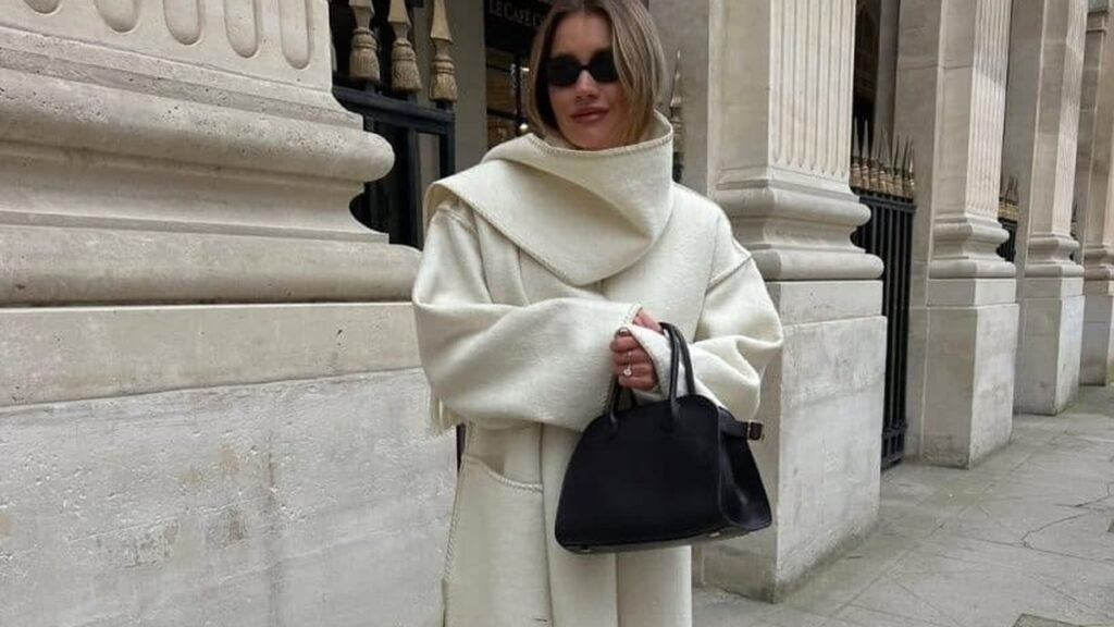 Woman wearing an oversized cream wool coat with a sculptural draped collar, holding a black leather handbag, standing against a stone colonnade in winter.