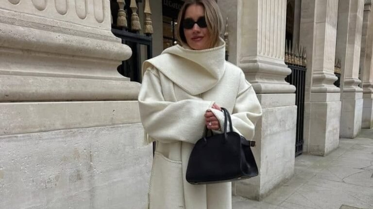 Woman wearing an oversized cream wool coat with a sculptural draped collar, holding a black leather handbag, standing against a stone colonnade in winter.