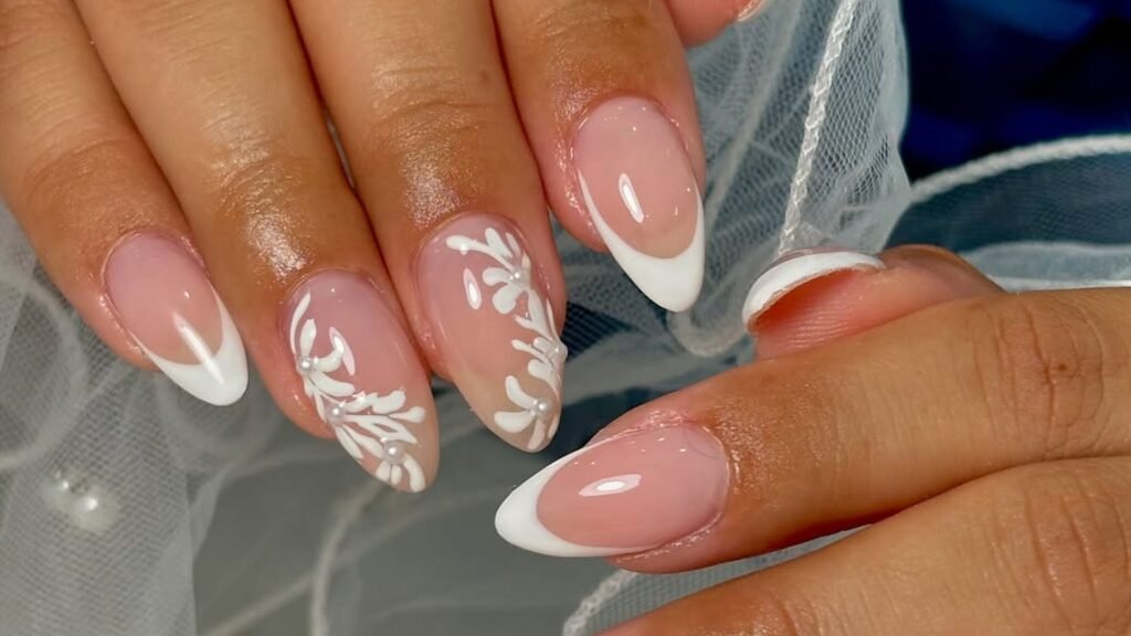 Close-up of two hands with almond-shaped blush nails, white French tips and hand-painted white leaf vines on two nails, resting against a sheer white bridal veil dotted with small pearls.