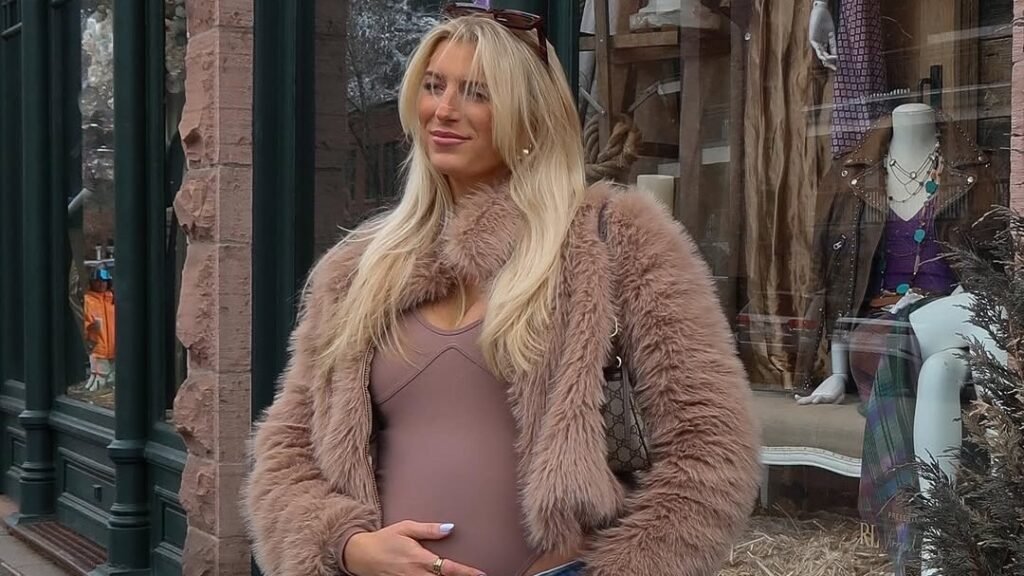 Pregnant woman in a fuzzy pink jacket and jeans standing outside a storefront, hand resting on her bump