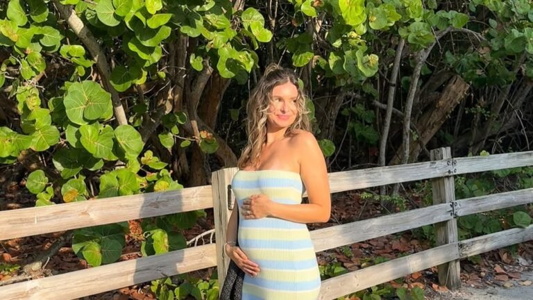 Pregnant woman in a fitted burgundy mesh dress cradling her bump against a brick wall