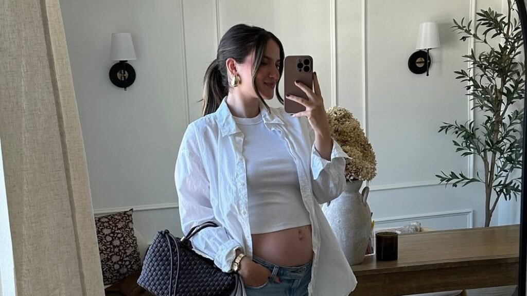 Pregnant woman in an oversized white button-down and cropped white tank, jeans unbuttoned and worn open under her bare bump, standing in a neutral living room