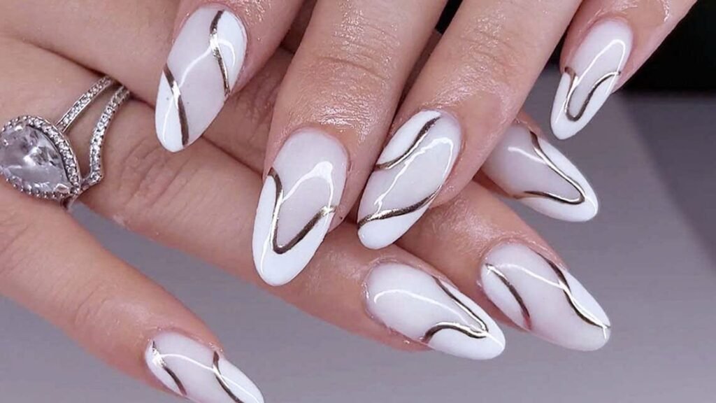 Hand with milky white almond-shaped bride nails decorated with thin metallic swirl lines, worn with a pear-shaped crystal ring against a soft gray background.