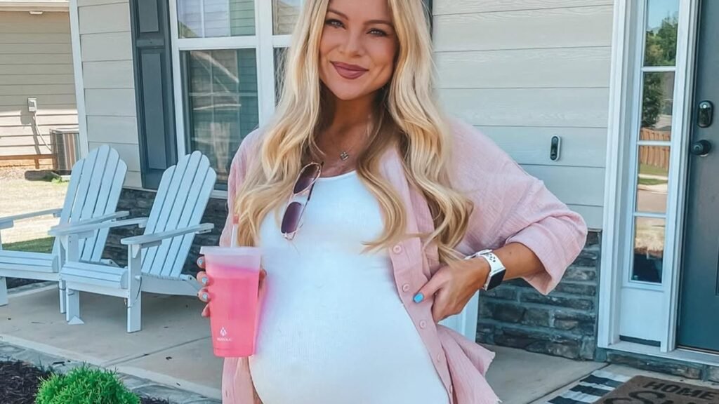 Smiling pregnant woman with long blonde hair in a white ribbed fitted dress and open pink gauze shirt, holding a pink tumbler while standing in front of blue porch chairs, a casual maternity outfit.