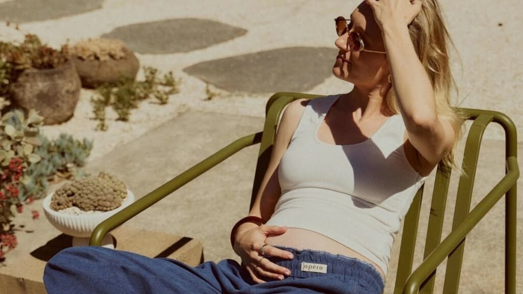 Pregnant woman relaxing outdoors in a white tank top and wide-leg denim pants with her bump exposed
