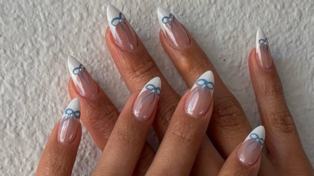 Almond-shaped nails with white French tips and hand-painted blue bows finished with tiny pearl centers, shown as a soft bridal shower nail idea