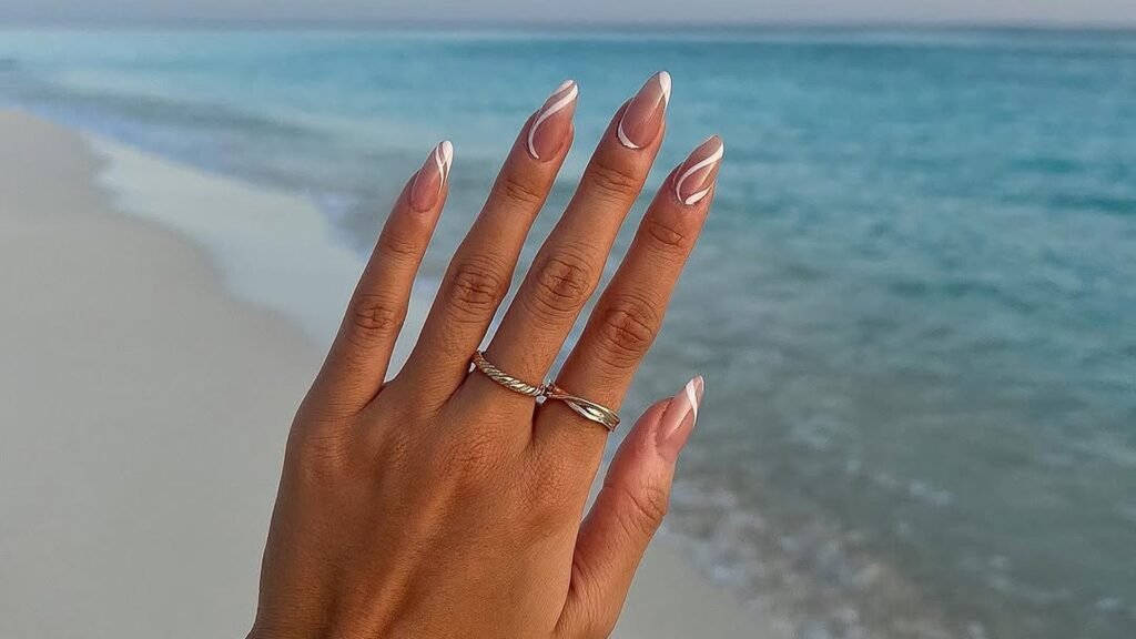 Hand raised in front of a calm turquoise ocean and pale sand shoreline, showing almond nails with white swirl French tips over a sheer nude base and two thin gold rings.