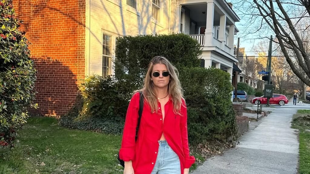 Woman wearing an oversized red button-down shirt open over light wash jeans, standing on a sidewalk in front of a red brick house at golden hour