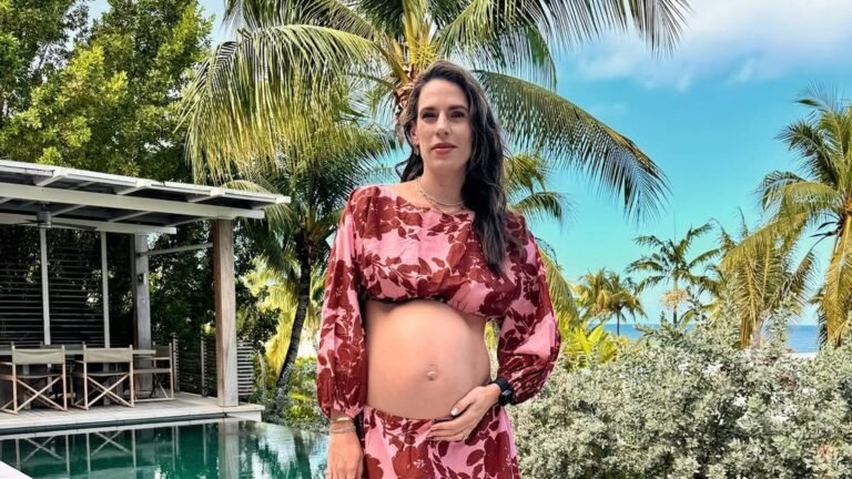 Pregnant woman in a pink and burgundy floral cropped top with balloon sleeves and a matching maxi skirt, her bare belly showing and one hand resting on it, standing beside a turquoise infinity pool under palm trees.